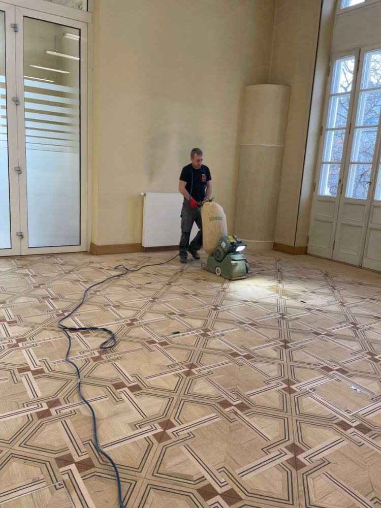 Zdjęcie prezentuje drewnianą podłogę pałacową budynku Uniwersytetu Warszawskiego, która poddawana jest zabiegowi renowacji wykonywanemu cykliniarką.