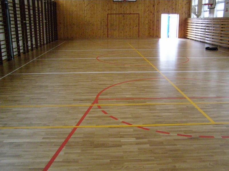 Zdjęcie przedstawia drewnianą podłogę sali gimnastycznej z profesjonalnie namalowanymi liniami boisk i zabezpieczoną lakierem.
